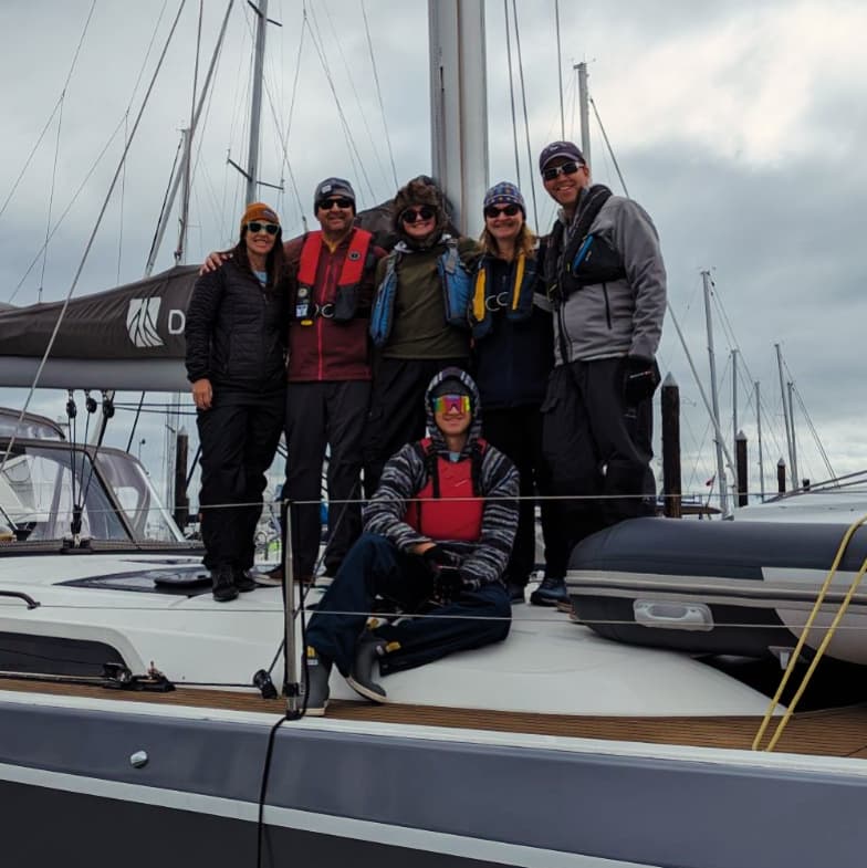 A group of six people posing on a sailboat in a marina, wearing outdoor clothing and life jackets, with cloudy skies in the background.